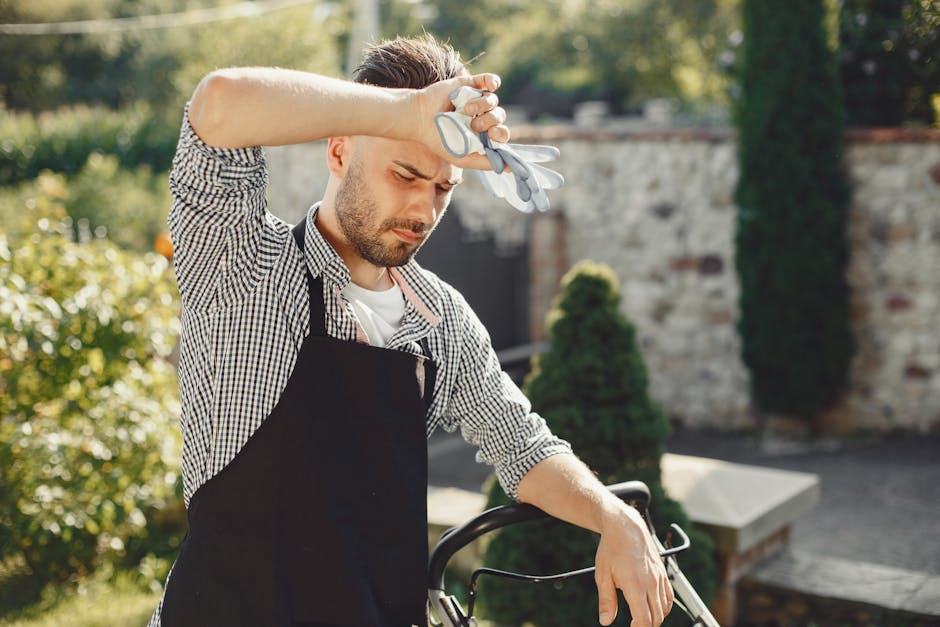A male gardener wearing a black apron over a checkered shirt stands outdoors on a sunny day, holding gardening gloves on his forehead with a slightly tired or contemplative expression. Behind him, the
